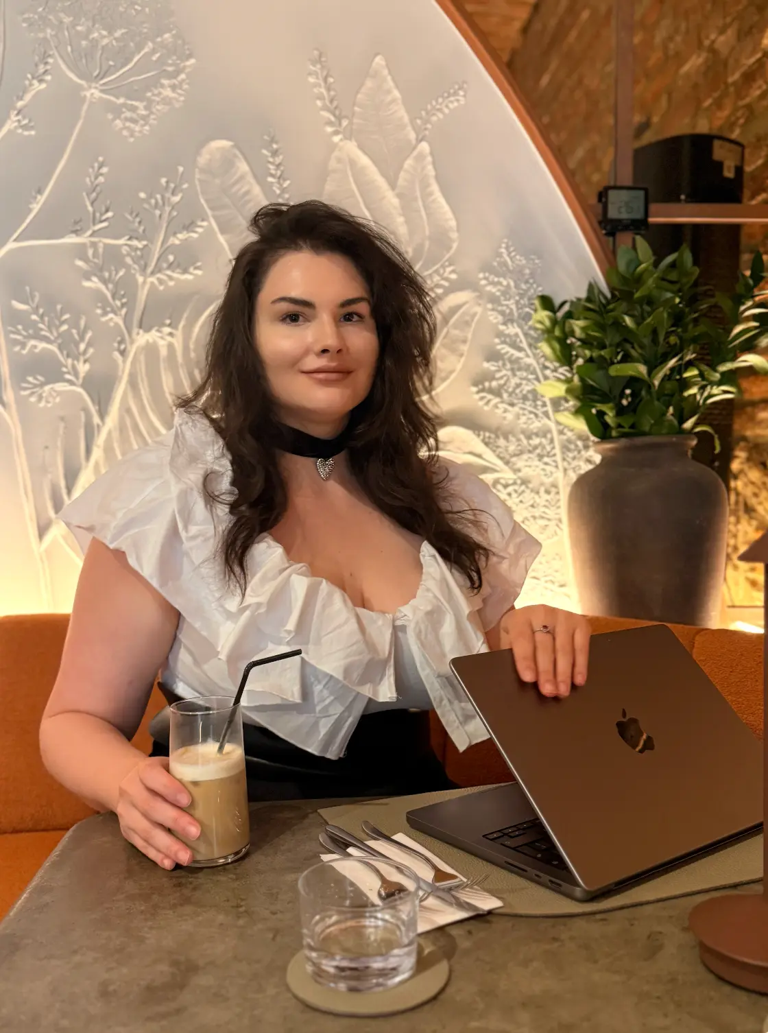 Andreea Chiș la masă, cu laptopul și o cafea cu paie, în fața unui perete cu motive florale